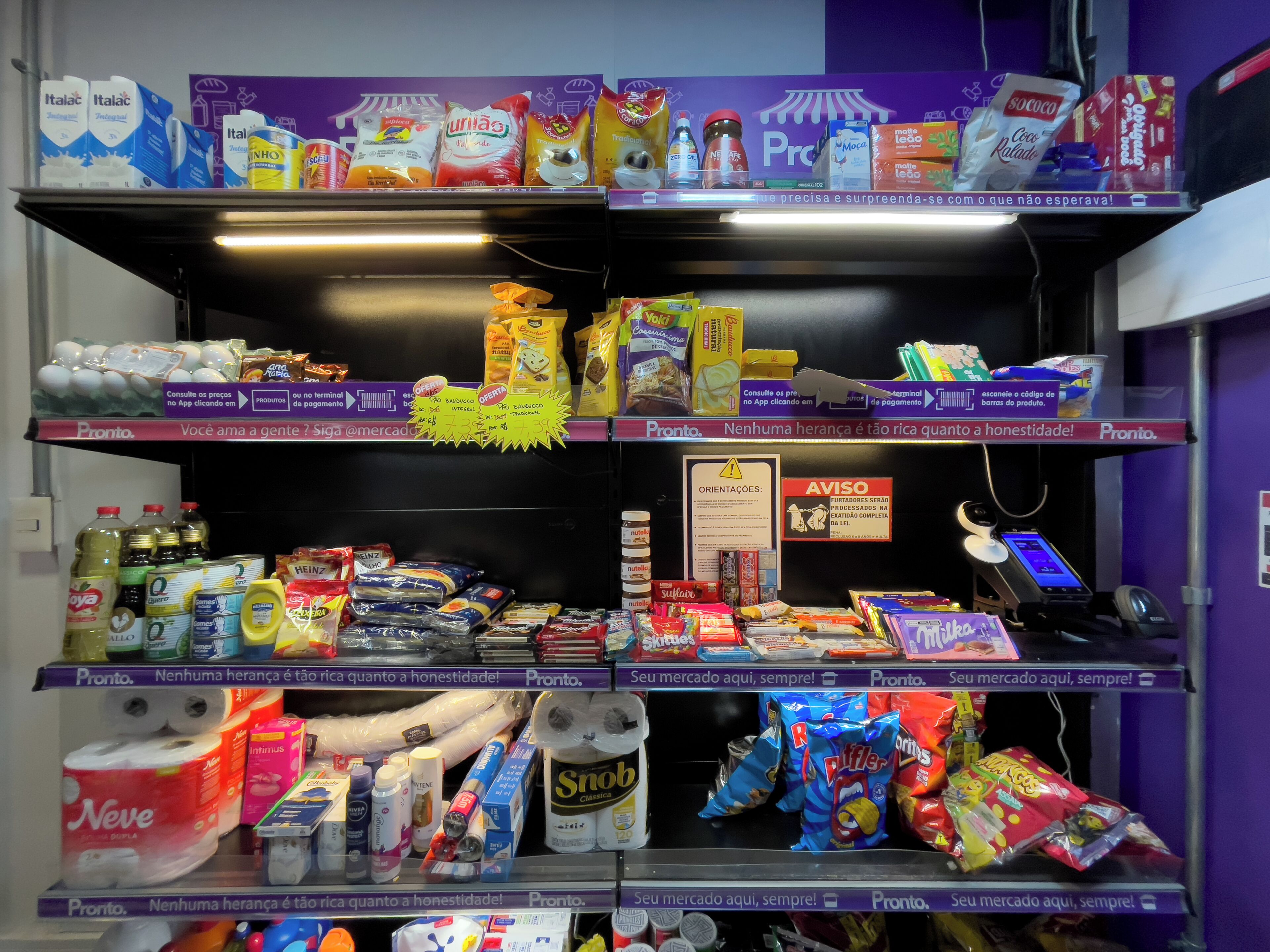 A self-service checkout area with various products on shelves, including snacks, beverages, and household items.