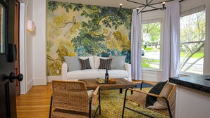 A living room with a white sofa, two wicker chairs, a coffee table, a mural of trees, and a view of the outdoors through a window. - Suite 1 at Kubli Haus - Luxury, Style & Location, 1 block to Britt (Jacksonville)
