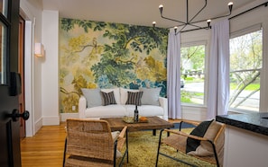 A living room with a white sofa, two wicker chairs, a coffee table, a mural of trees, and a view of the outdoors through a window.
