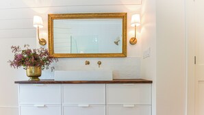 A bathroom with a white vanity, gold-framed mirror, two wall-mounted lights, and a potted plant. - Suite 4 at Kubli Haus<br>Luxury, Style & Location, 1 block to Britt (Jacksonville)