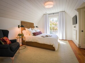 Un dormitorio con una cama grande, una silla, una mesita con una planta, un televisor montado en la pared y una ventana con cortinas.