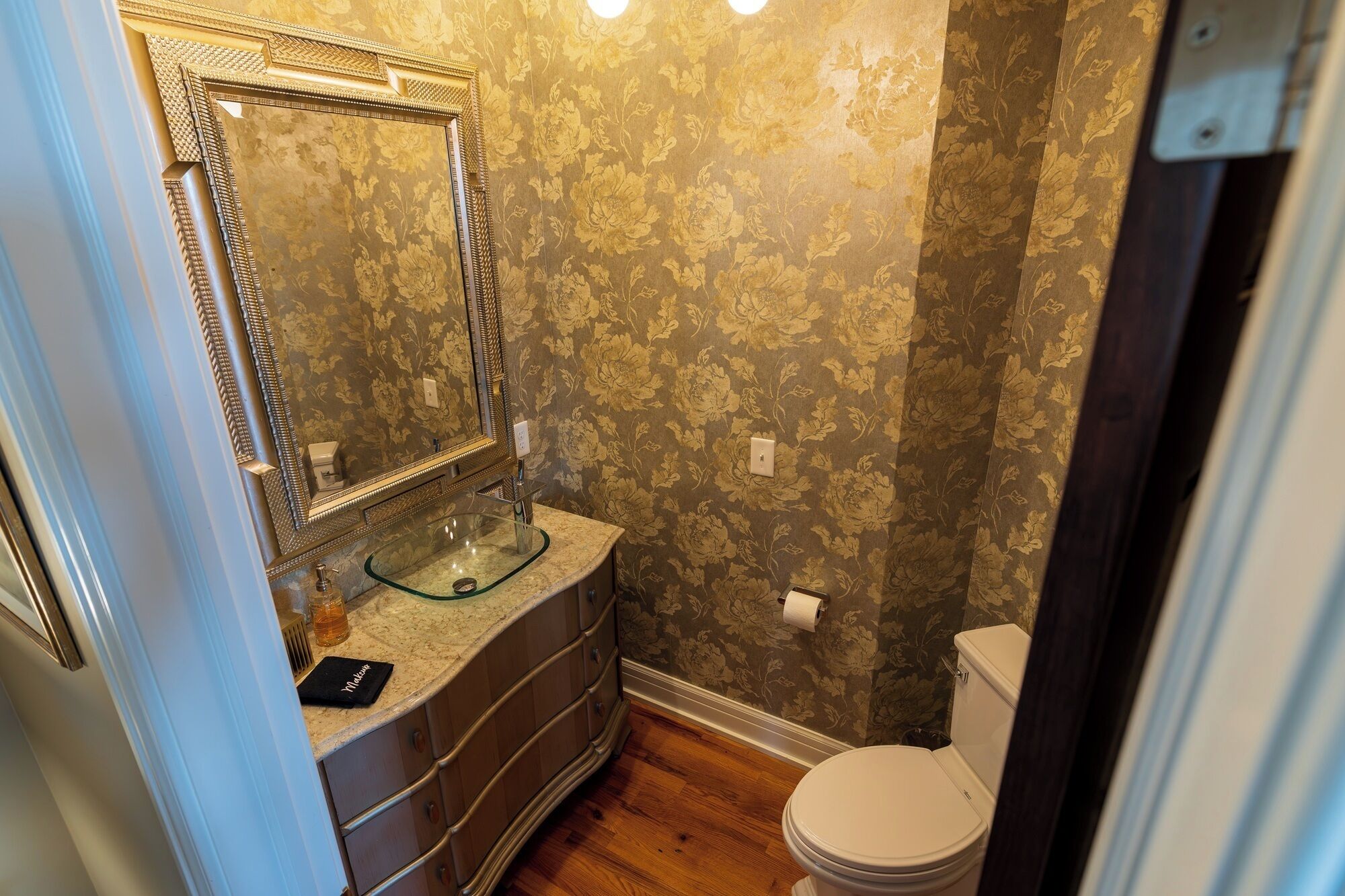 Un bany amb un lavabo de vidre, un mirall gran, un vàter i paper pintat amb motius florals.