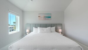 A bedroom with a large bed, two bedside tables with lamps, a painting above the bed, and a window with a view. - Shore Bet (Fort Myers Beach)