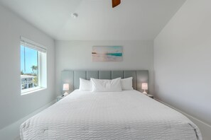 A bedroom with a large bed, two bedside tables with lamps, a painting above the bed, and a window with a view. - Shore Bet (Fort Myers Beach)
