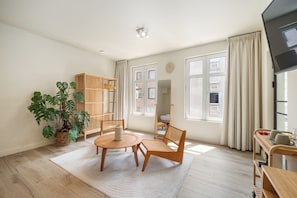 Una sala de estar moderna con piso de madera, una mesa redonda, dos sillas, una planta grande en maceta, una estantería de madera y un ventanal con cortinas.