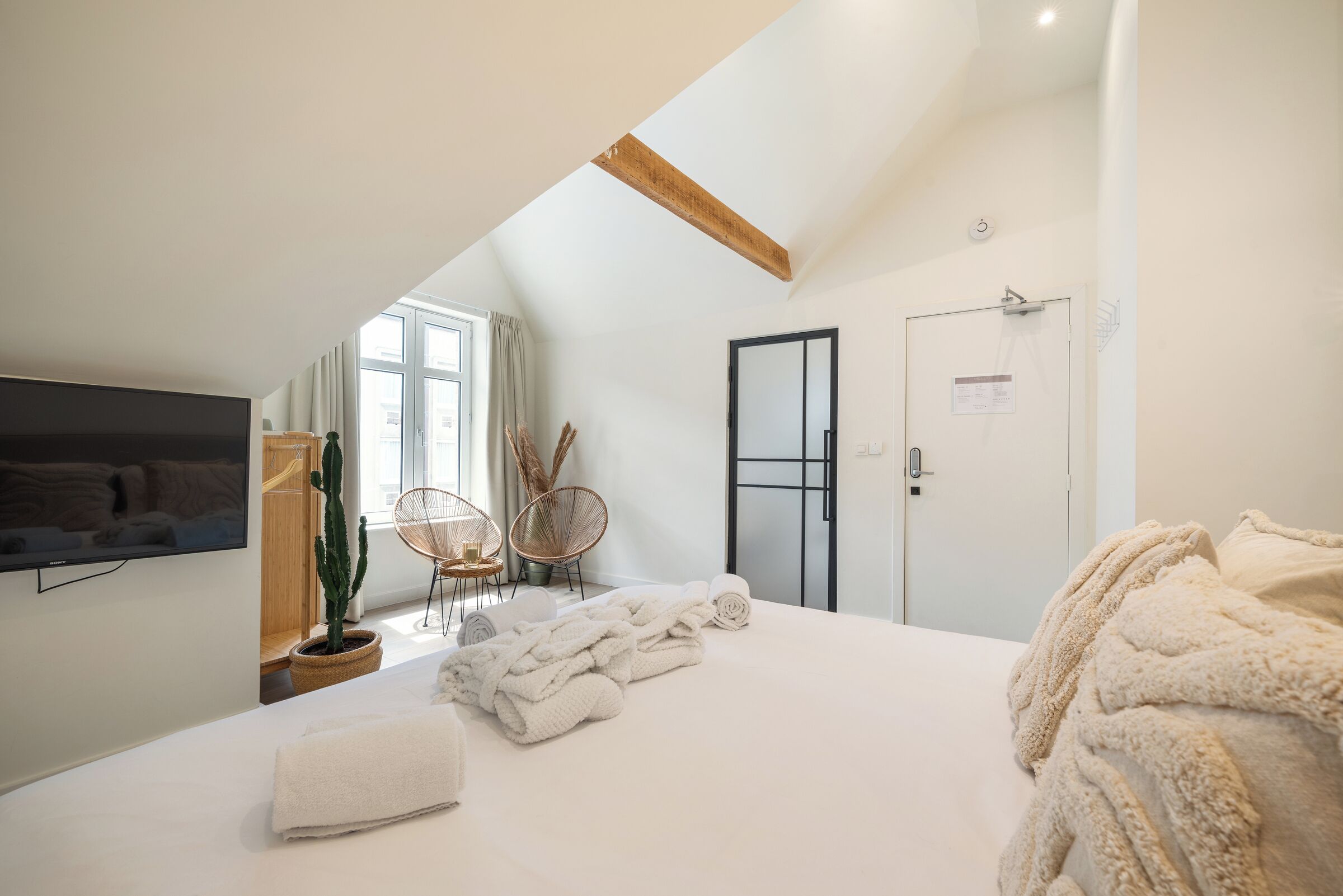 A bedroom with a sloped ceiling, a bed with white linens, a television mounted on the wall, and a door with a sign.