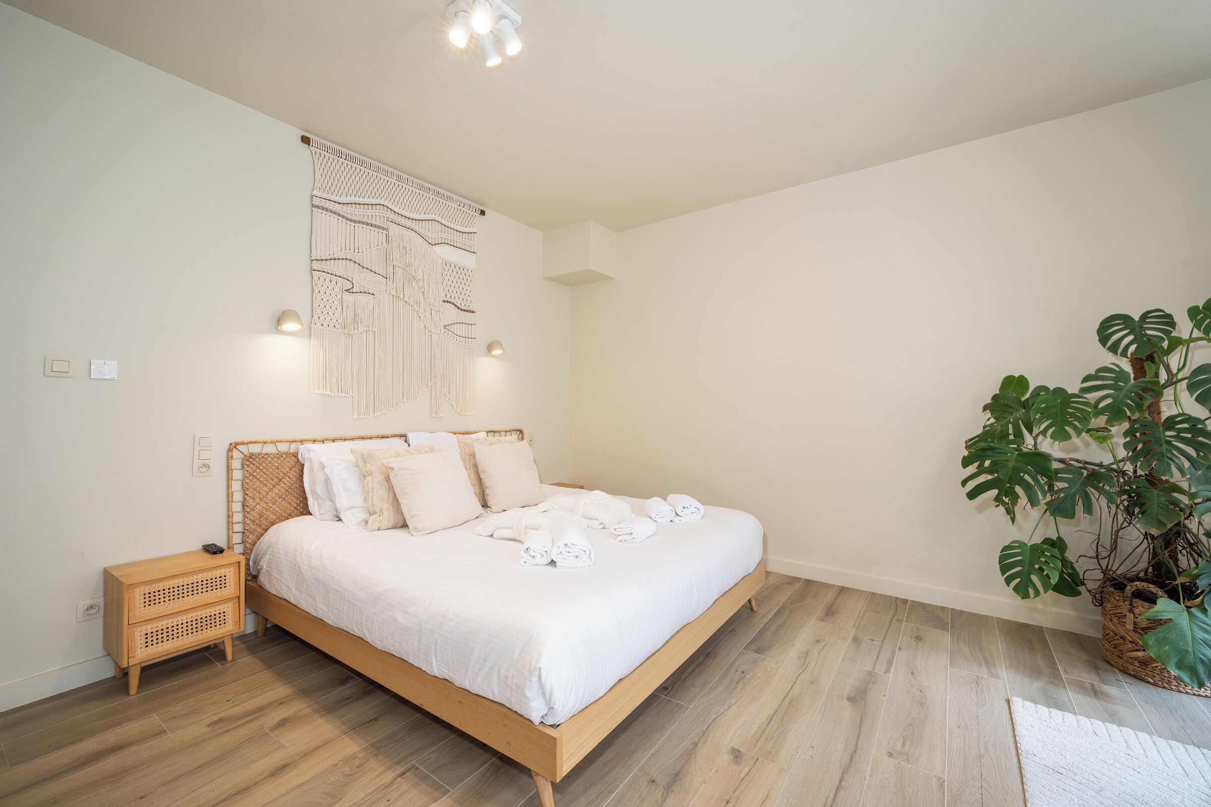 Een net opgemaakt bed met witte lakens, een nachtkastje, een wandlamp en een potplant.