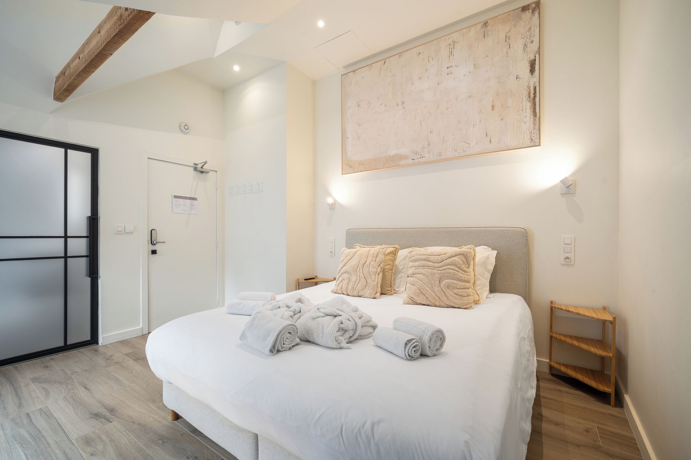 Une chambre d’hôtel moderne avec un grand lit, une poutre en bois au plafond, une œuvre d’art accrochée au mur et une petite table d’appoint en bois.