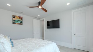 A bedroom with a bed, a ceiling fan, a TV mounted on the wall, and a door. - Beach Please (Fort Myers Beach)