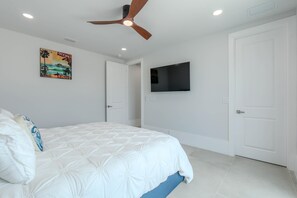 A bedroom with a bed, a ceiling fan, a TV mounted on the wall, and a door. - Beach Please (Fort Myers Beach)