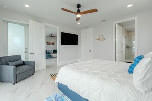 Ein Schlafzimmer mit Bett, grauem Sofa, Deckenventilator, Fernseher und einer Badezimmertür.