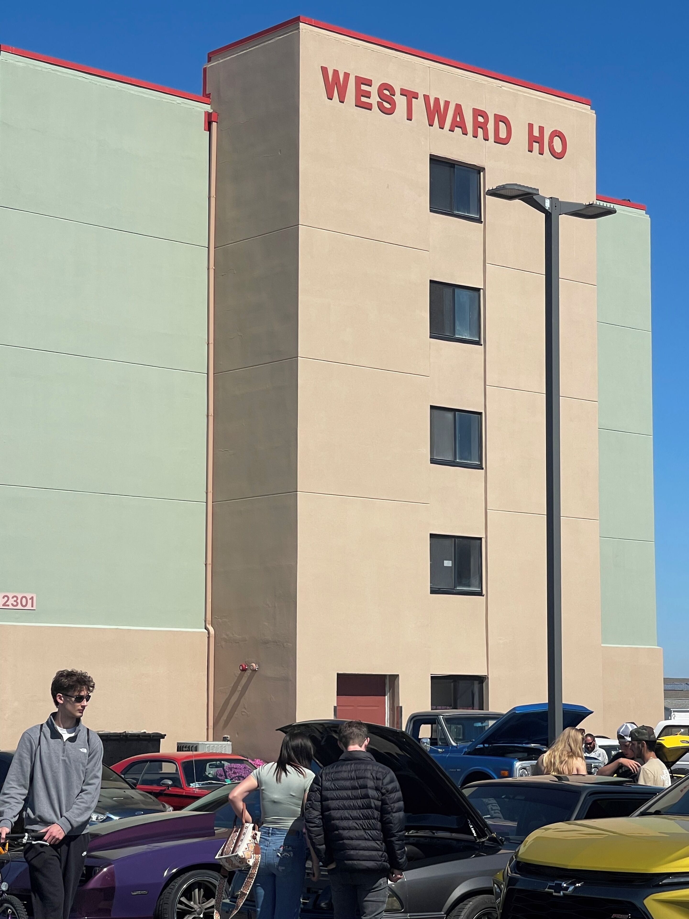 A building with 'WESTWARD HO' signage, a parking lot with various cars, and a clear blue sky.