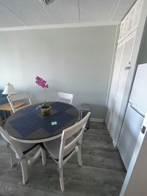 A small, compact kitchen with a dining table, chairs, a trash bin, and a refrigerator. - Cute 1-bedroom apt 23rd St in Ocean City near the beach (Ocean City)