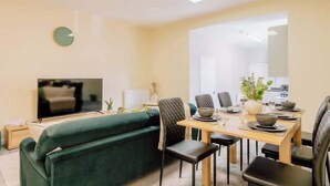 A living room with a TV, green sofa, dining table with chairs, and a clock on the wall. - Contractor and Business Stays | Sleeps 6 | WiFi (Hertfordshire)