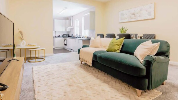 A living room with a green sofa, a TV, a small table with a plant, and a kitchen in the background. - Contractor and Business Stays | Sleeps 6 | WiFi (Hertfordshire)