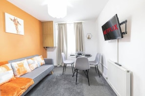 A living room with a sofa, dining table, chairs, a TV mounted on the wall, and a radiator. - Contractor-Friendly | Secure Parking & Fast Wi-Fi (West Midlands)