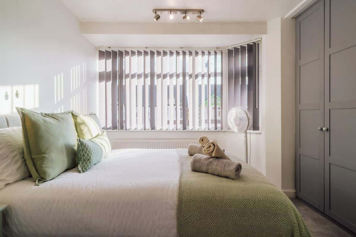 Un dormitorio con cama, almohadas, un radiador, una ventana con persianas, un ventilador y un armario.