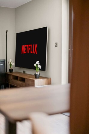A modern living room with a flat-screen TV mounted on the wall displaying 'NETFLIX,' a wooden coffee table, and two potted plants. - Sleeps 6 | Relocation & Long Stays, FREE Parking (Greater London)