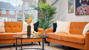 Un salon avec deux canapés orange, une table basse en verre, une grande plante et un tableau accroché au mur.