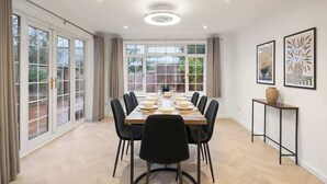 Une salle à manger avec une grande table dressée pour six personnes, entourée de chaises, et offrant une vue sur l’extérieur grâce à de grandes fenêtres.
