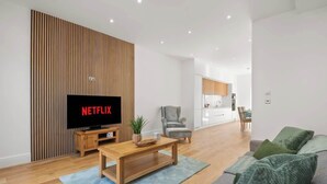 Un salon moderne avec un téléviseur à écran plat, un mur recouvert de panneaux en bois, un canapé gris et une cuisine en arrière-plan.