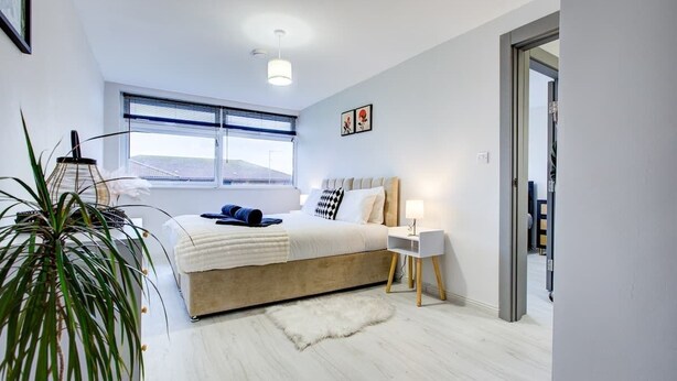 Une chambre à coucher moderne avec un grand lit, une table de chevet, une lampe, une plante et une vue sur l’extérieur.