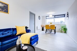 Un salón moderno con un sofá azul, cojines amarillos, un área de comedor con sillas amarillas y un ventanal con vistas.