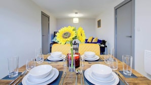 Une table à manger dressée pour quatre personnes, avec un vase de tournesols, une bougie, et un lit avec une tête de lit jaune en arrière-plan.