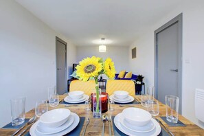 Mesa de comedor dispuesta para cuatro personas, con un jarrón de girasoles, una vela y una cama con cabecero amarillo al fondo.