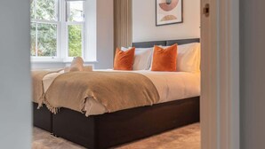 Une chambre avec une grande fenêtre, un lit avec des coussins orange et un plaid beige.