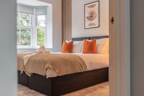Ein Schlafzimmer mit großem Fenster, einem Bett mit orangefarbenen Kissen und einer beigen Tagesdecke.