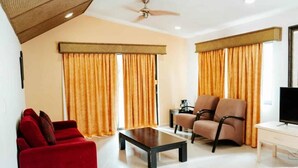 A hotel room with a red sofa, a black coffee table, a beige armchair, a TV stand, and a bed with a patterned bedspread.