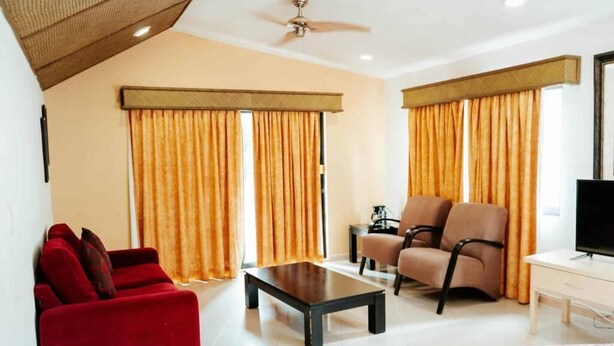 Habitación de hotel con un sofá rojo, una mesa de centro negra, un sillón beige, un mueble para televisor y una cama con colcha estampada.