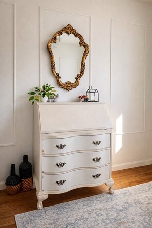 A white dresser with ornate gold trim, a mirror with a gold frame, and a small plant on top.