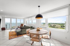 A modern living room with a round dining table, two chairs, a sofa, and a balcony with a view. - Santa Rosa Dunes 1061 (Pensacola Beach)