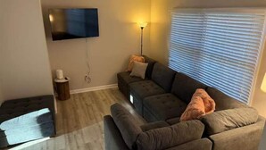 Una sala de estar con un sofá modular, un televisor de pantalla plana montado en la pared, una lámpara de pie y una ventana con persianas.