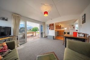 A living room with a TV, green sofas, a dining area, and a kitchen with a refrigerator and shelves. - Dragon Fly Cottage - Te Anau Holiday Home (Te Anau)