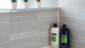 Casa de banho com paredes revestidas a azulejos cinza, uma janela com porta e duas embalagens de produtos TRESemmé sobre o balcão.