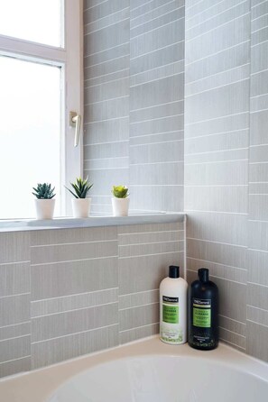 A bathroom with grey tiled walls, a window with a door, and two bottles of TRESemmé products on the counter.