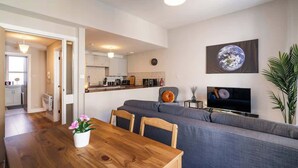 Uma sala de estar moderna com uma mesa de jantar de madeira, sofá cinzento e uma televisão de tela plana na parede.