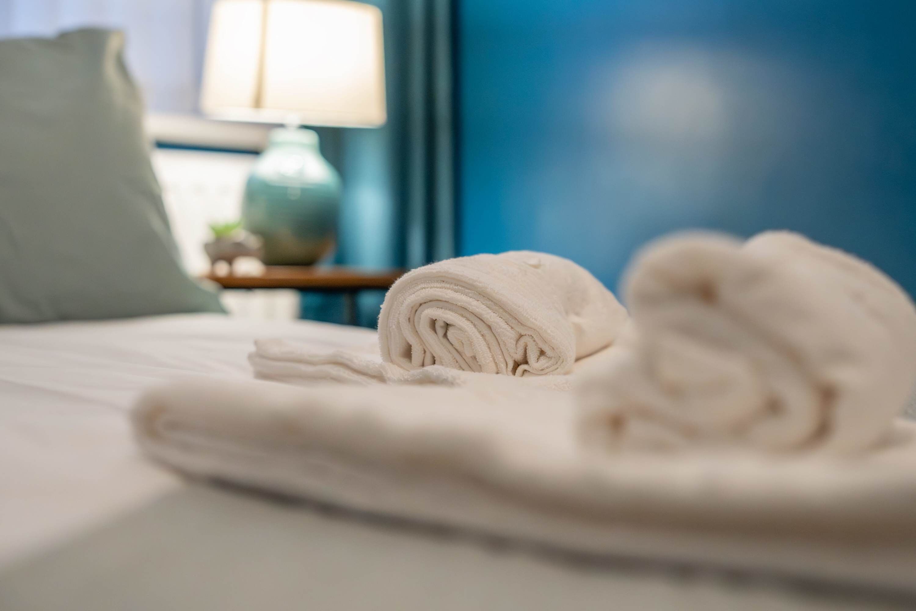A neatly made bed with rolled towels and a pillow, a lamp on a side table, and a blue wall in the background.