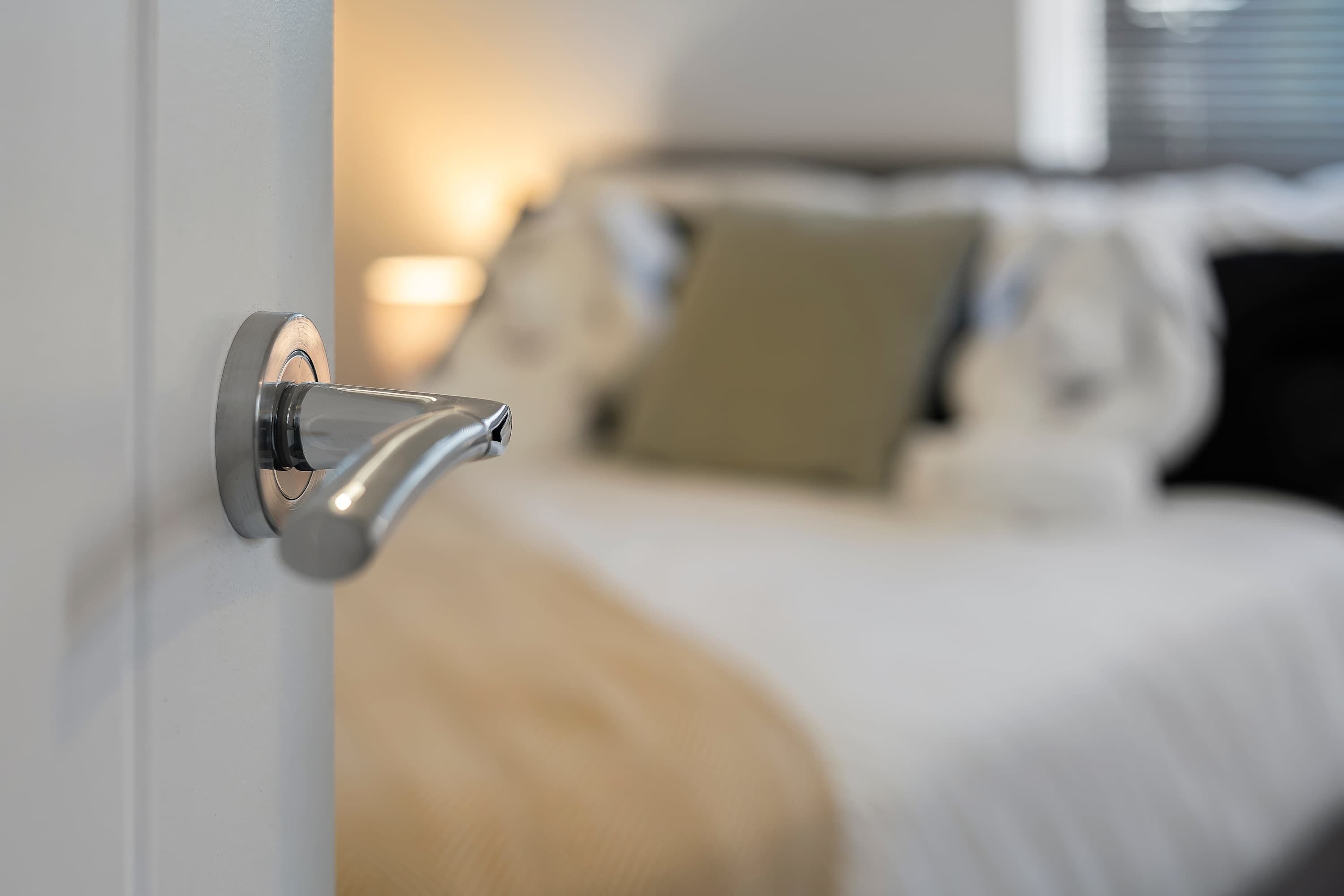 Une chambre d’hôtel moderne avec une poignée de porte métallique, un lit recouvert d’une couette blanche et beige, et une lampe de chevet.