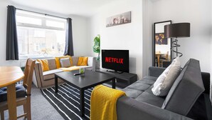 Ett modernt vardagsrum med en soffa, ett soffbord, en tv som visar "NETFLIX", en lampa och utsikt över omgivningen.