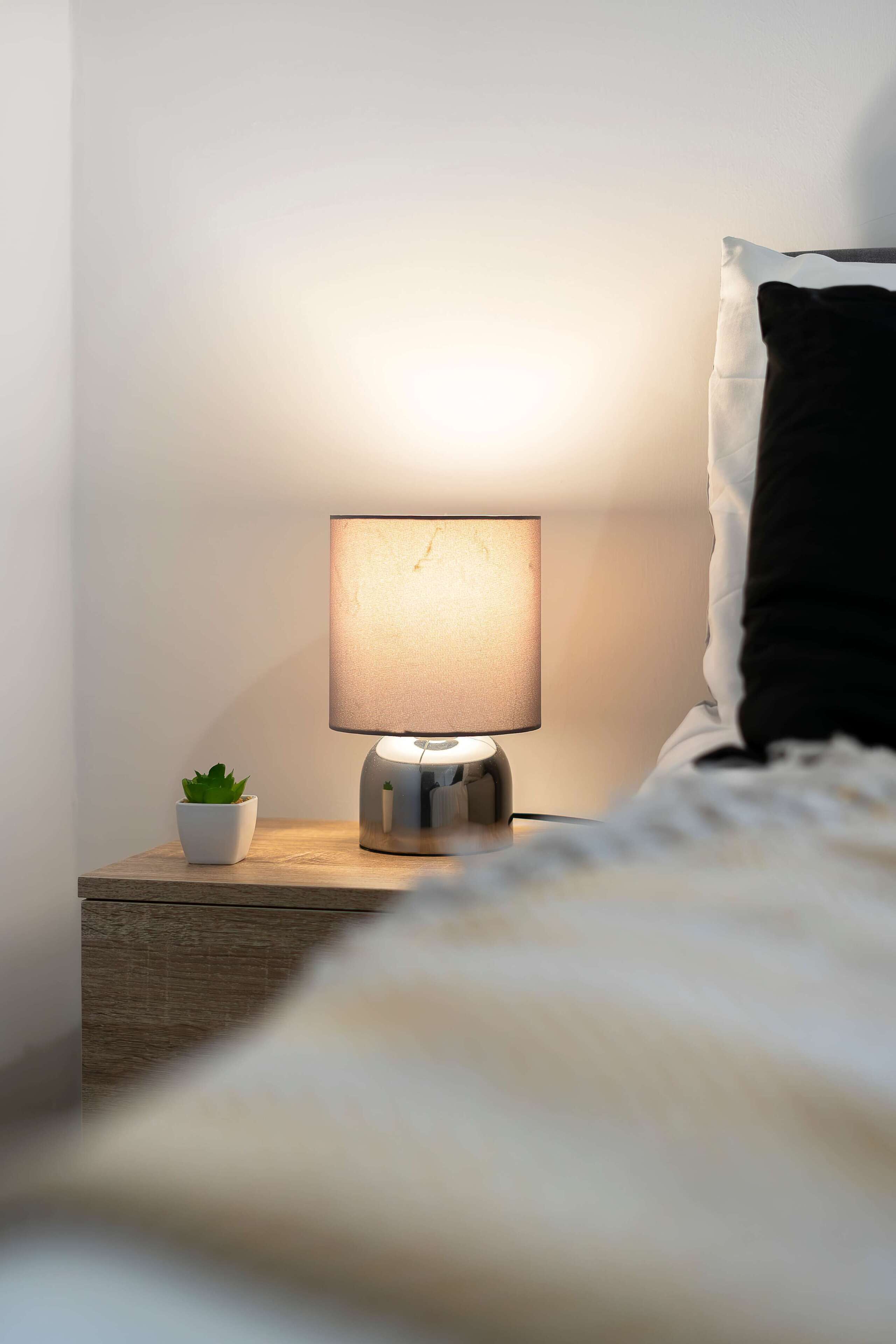 A bedside table with a lamp, a small potted plant, and a black pillow on a bed.