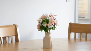 Ett runt träbord med en vas med blommor, två trästolar och en enkel, vit vägg med ett fönster.