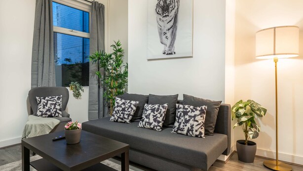 A living room with a sofa, a coffee table, a floor lamp, a potted plant, and a window with curtains. - 10 Mins BAE Systems -Contractor Stay -Free Parking (Barrow-in-Furness)