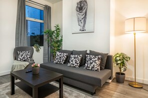 Una sala de estar con un sofá, una mesa de centro, una lámpara de pie, una planta en maceta y una ventana con cortinas.