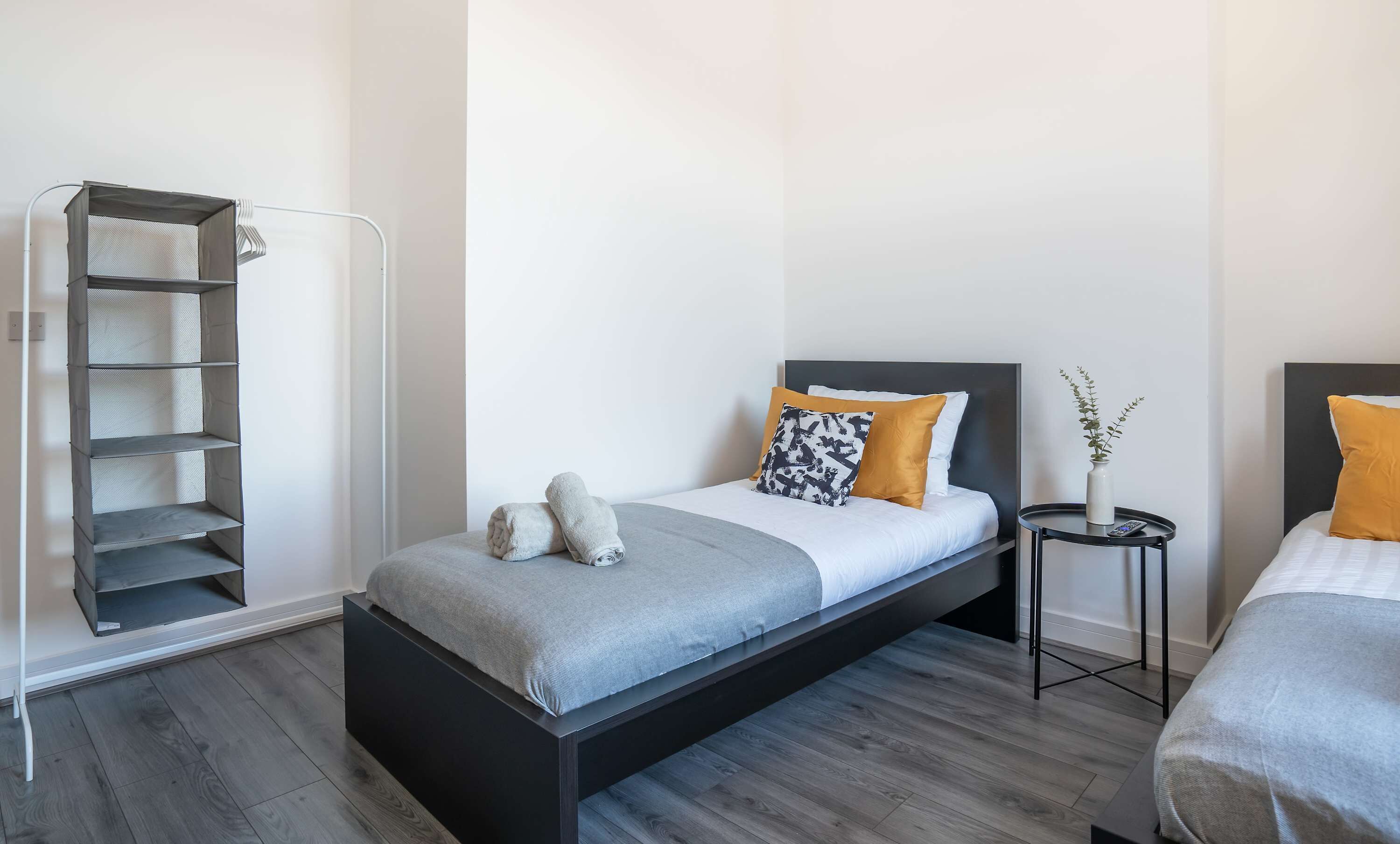 A modern bedroom with a bed, a side table, a small shelf unit, and a bedside plant.