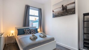 A bedroom with a bed, a window with curtains, a radiator, a bedside table with a lamp, and a wall-mounted shelf. - 10 Mins BAE Systems -Contractor Stay -Free Parking (Barrow-in-Furness)