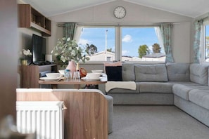 A cozy living area with a sofa, a dining table with dishes, a clock, and a view through the window. - Seton Sands 3BR Spacious Caravan near Edinburgh (Port Seton)